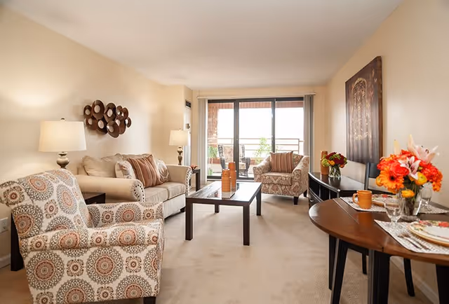 A cozy living room with beige walls and carpet, featuring a patterned armchair, a beige sofa with striped pillows, a wooden coffee table, and a side table with a lamp. There is a sliding glass door leading to a balcony with outdoor seating. A round dining table with flowers and place settings is visible on the right side of the room.