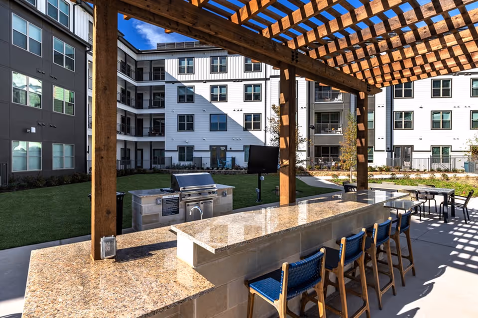 Outdoor courtyard with a wooden pergola, stone bar counter and blue bar stools, a grill, and surrounding multi-story apartment building.