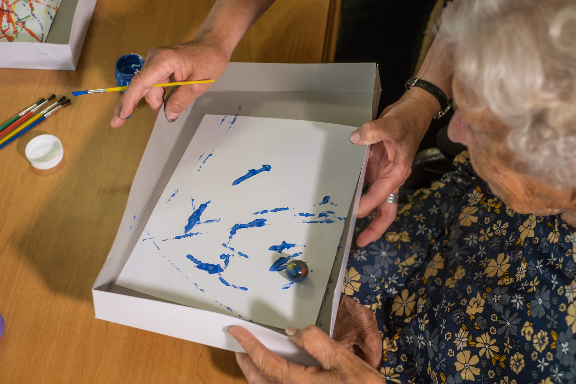 An elderly woman and another person are engaged in a painting activity using a marble and blue paint on a white sheet inside a shallow box on a wooden table. Several paintbrushes and paint containers are visible nearby.
