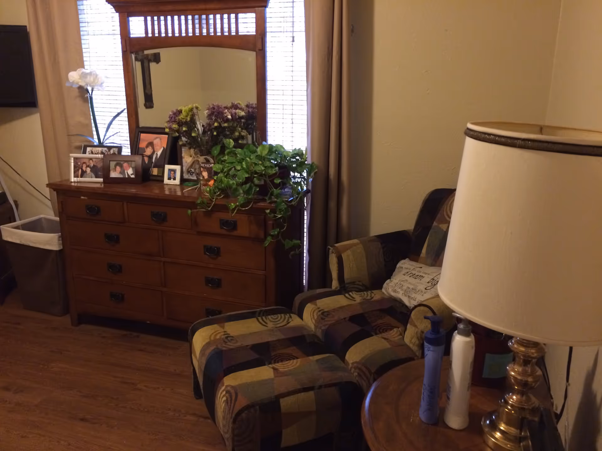A cozy interior room featuring a wooden dresser with a large mirror, decorated with framed photos, flowers, and a green plant. Next to the dresser is a patterned armchair with a matching ottoman and a pillow. A round wooden side table with a brass lamp and two lotion bottles is also visible. The room has wooden flooring and beige walls with a window covered by blinds and curtains behind the dresser.