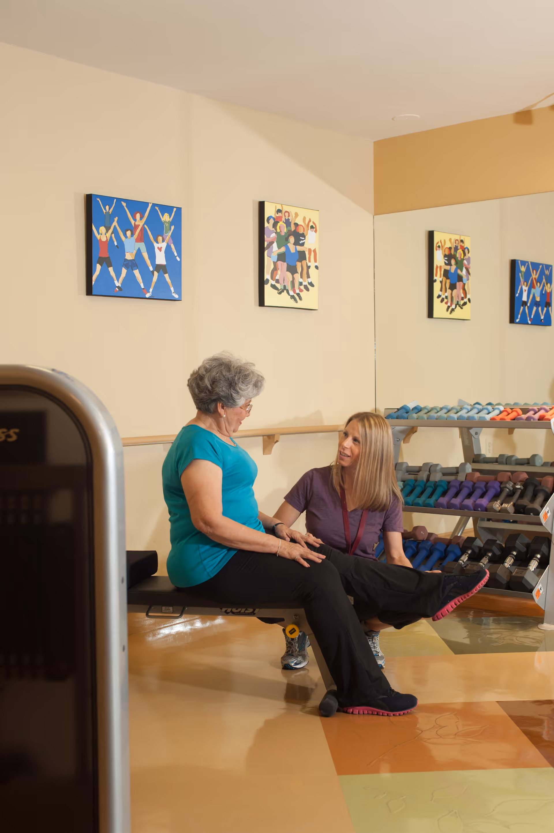 An elderly woman in a turquoise shirt and black pants is seated on a workout bench while a fitness instructor in a purple shirt assists her with leg exercises in a gym room. The room has a rack of colorful dumbbells and two paintings of people exercising on the wall.