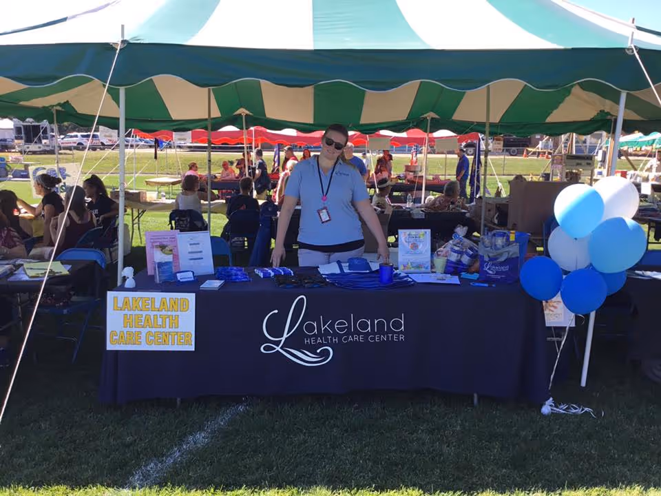 A woman standing behind a table covered with a black tablecloth that has the Lakeland Health Care Center logo. The table is set up under a green and white striped tent at an outdoor event. There are informational materials and blue and white balloons on the right side of the table. People are seated and walking around in the background on the grass.