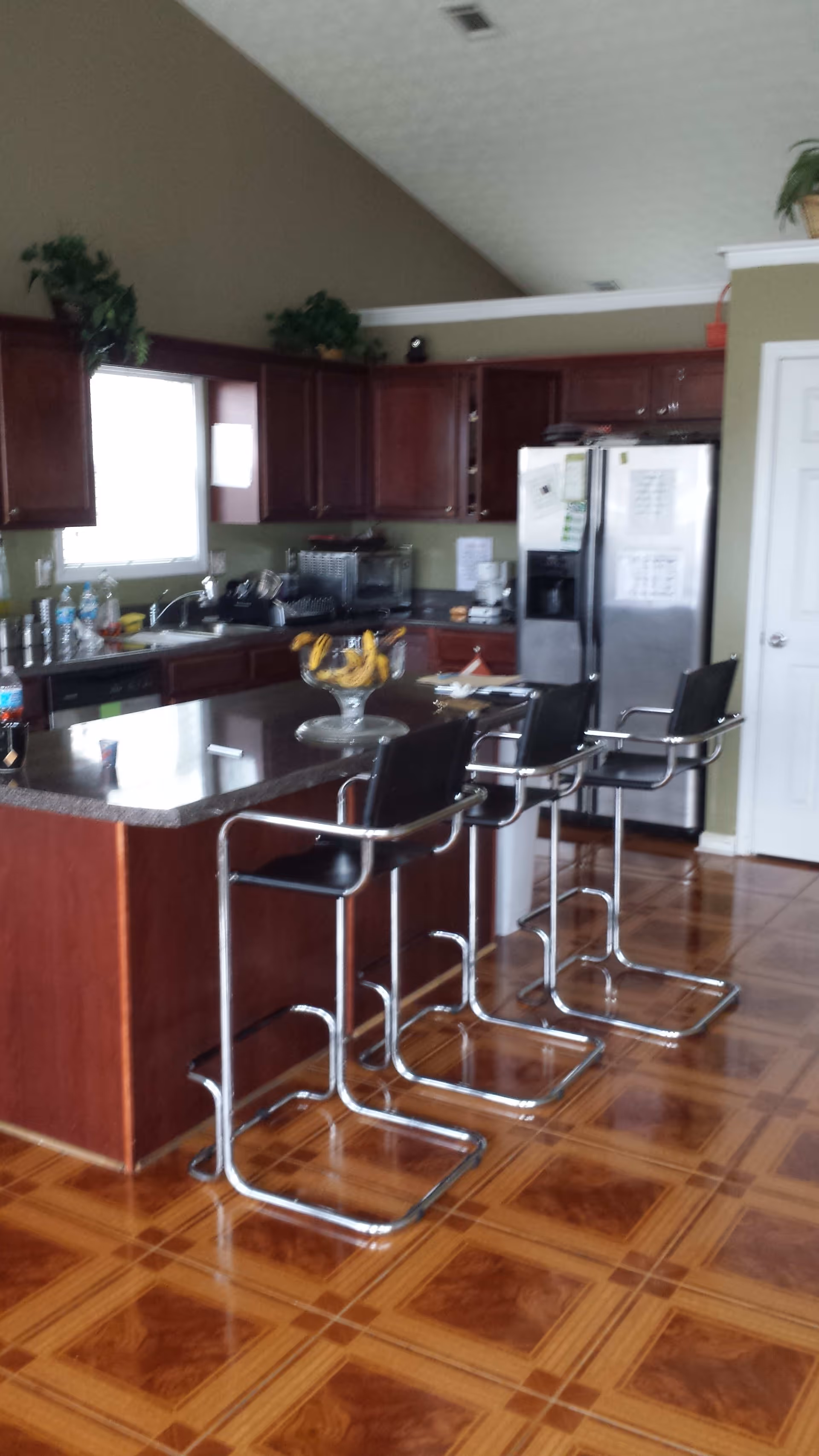 A kitchen with wooden cabinets, a stainless steel refrigerator, a countertop island with three modern bar stools, and a bowl of bananas on the island. The floor has a patterned wood design, and there are plants on top of the cabinets.