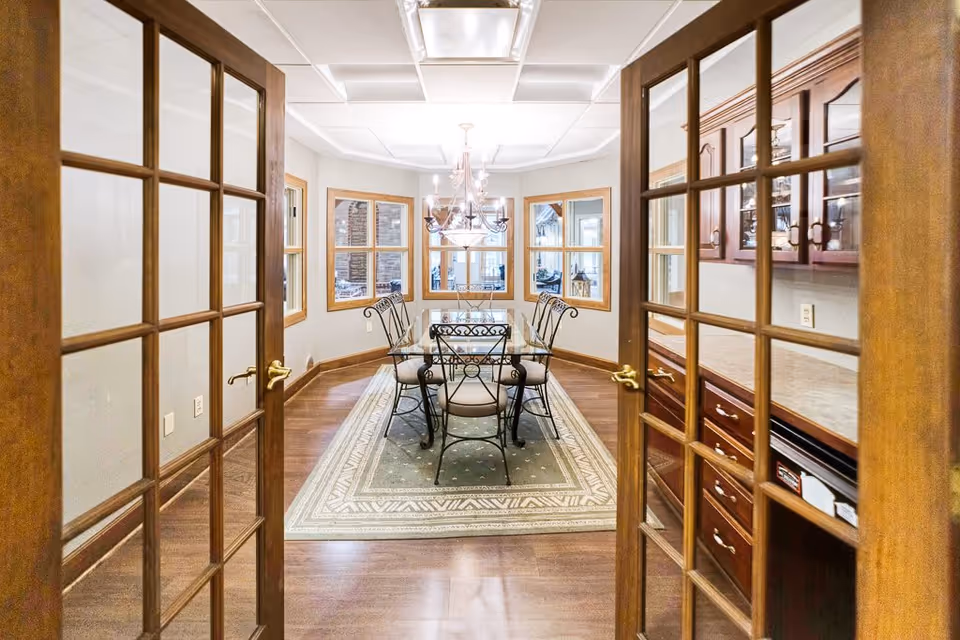 View through wooden framed glass double doors into a dining room with a glass-top table and six wrought iron chairs on a patterned area rug. The room has hardwood floors, a chandelier hanging from the ceiling, and multiple windows with wooden frames.