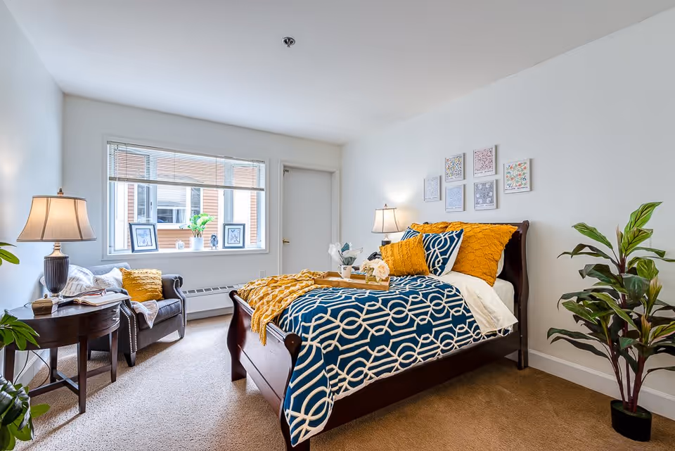 A bright and cozy bedroom with a large window letting in natural light. The room features a wooden bed with blue and white patterned bedding and mustard yellow pillows and throw blanket. There is a small round side table with a lamp and a comfortable armchair with a yellow pillow and a blanket. Several framed pictures hang on the white wall above the bed, and a green potted plant is placed in the corner near the bed.