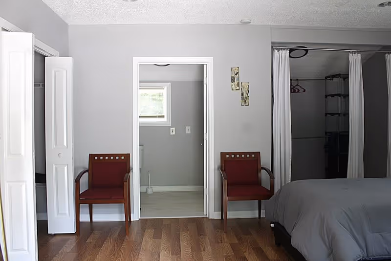 Simple bedroom interior showing two chairs flanking a central doorway, a bed on the right, and open closets.