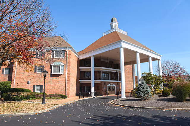 Exterior view of a senior living facility named Richmond Place, featuring a large brick building with a covered entrance supported by white columns, surrounded by landscaped bushes and trees with autumn leaves.