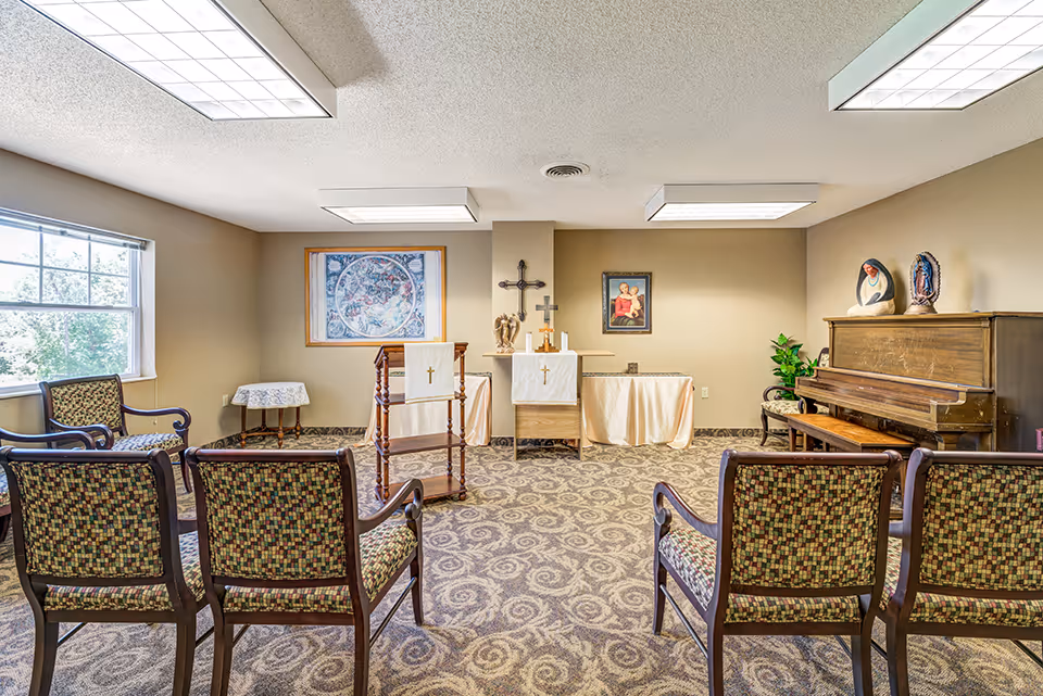 A small chapel room with several chairs arranged in rows facing an altar. The altar is adorned with white cloths, crosses, and religious statues. There is a piano on the right side of the room with religious figurines on top. The room has beige walls, a patterned carpet, and fluorescent ceiling lights. A window on the left side lets in natural light.