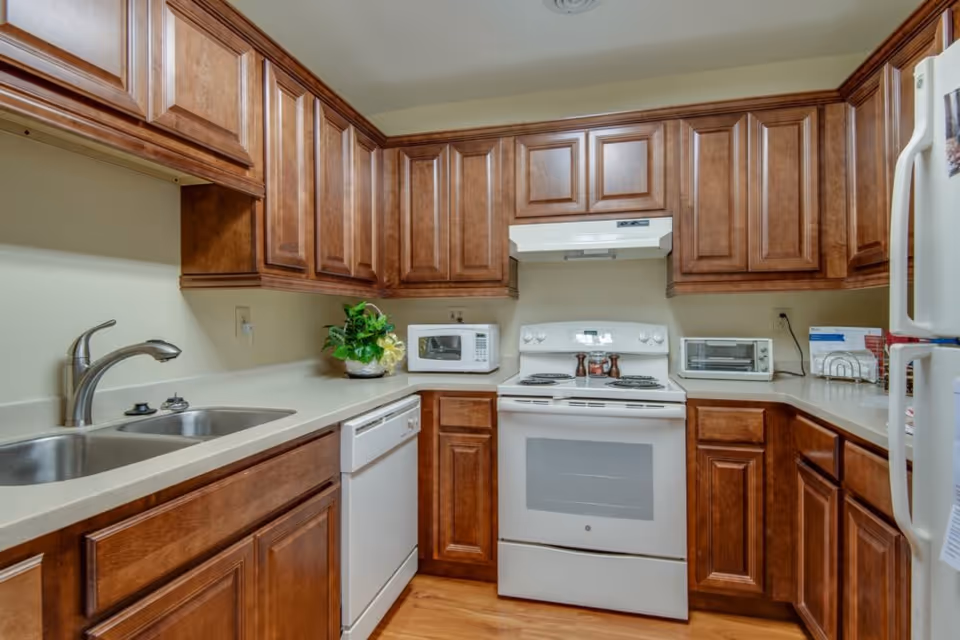 Compact U-shaped kitchen featuring wooden cabinets, a white stove and dishwasher, double sink, microwave, toaster oven, and refrigerator.