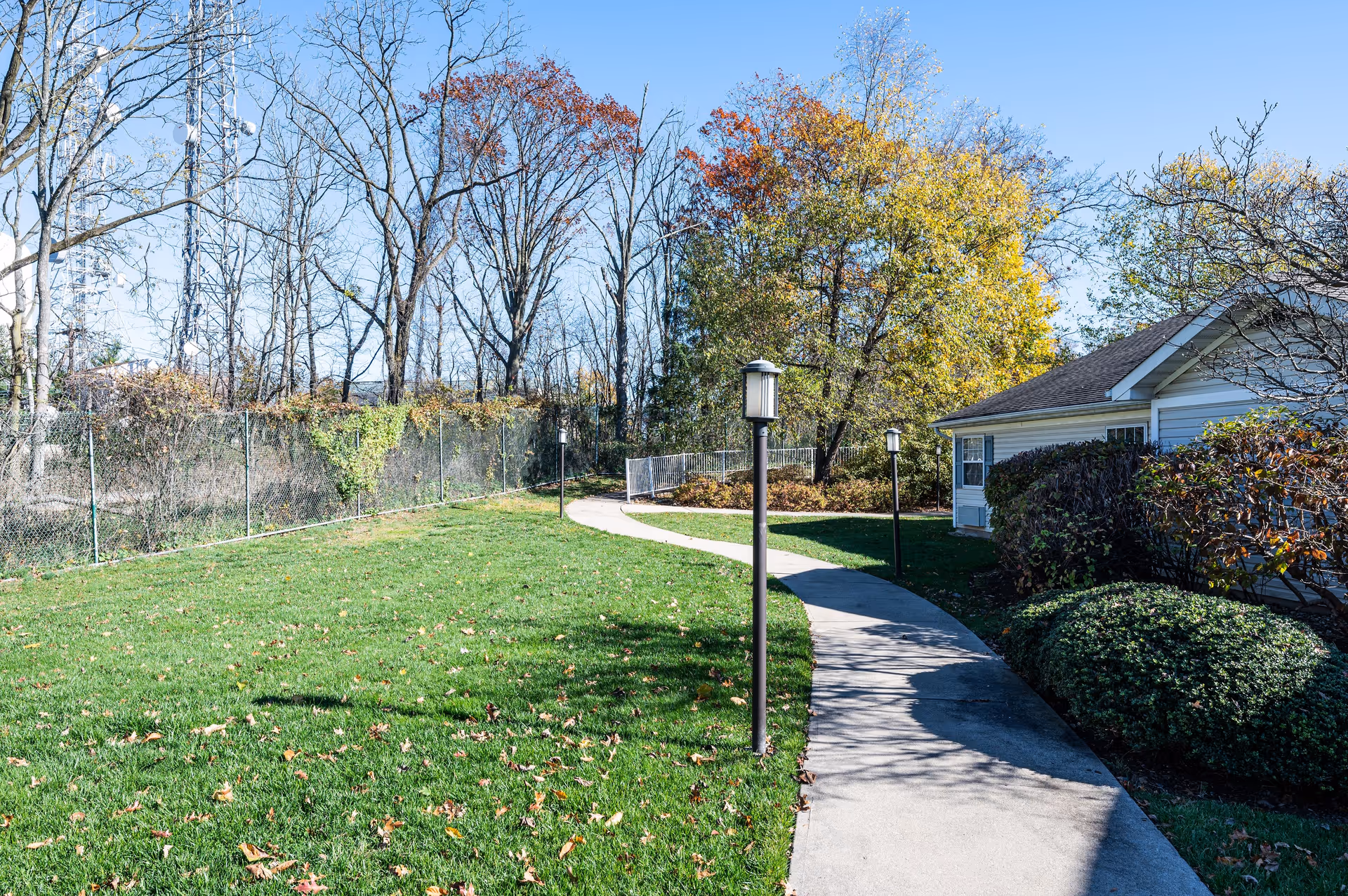 A winding concrete pathway bordered by green grass and small lamp posts leads past a white building with bushes and trees showing autumn foliage under a clear blue sky.