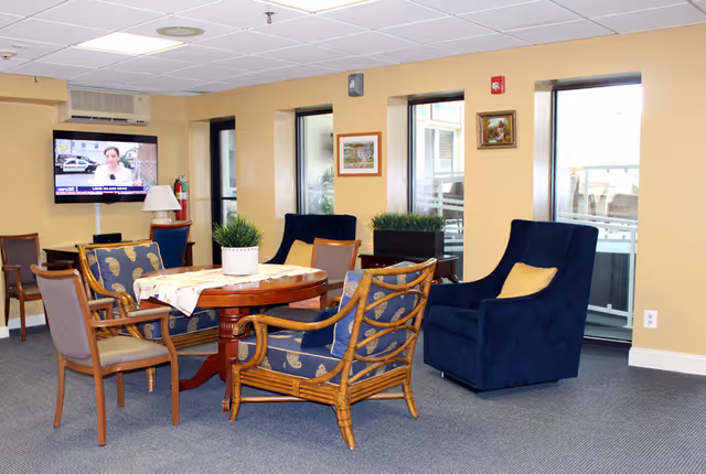 A cozy common area in an assisted living facility with a round wooden table surrounded by various chairs, including cushioned armchairs and wooden chairs. The room has beige walls, three windows with views outside, framed artwork on the walls, and a flat-screen TV mounted on the wall showing a news broadcast. There are potted plants on the table and near the windows, and the floor is carpeted in gray.