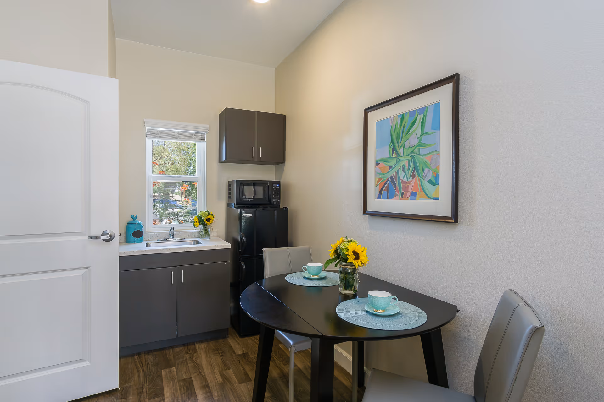 Small kitchen area with a black round table set for two with teal placemats, cups, and a vase of sunflowers. There is a black mini refrigerator with a microwave on top, gray cabinets, a sink under a window, and a colorful framed painting on the wall.