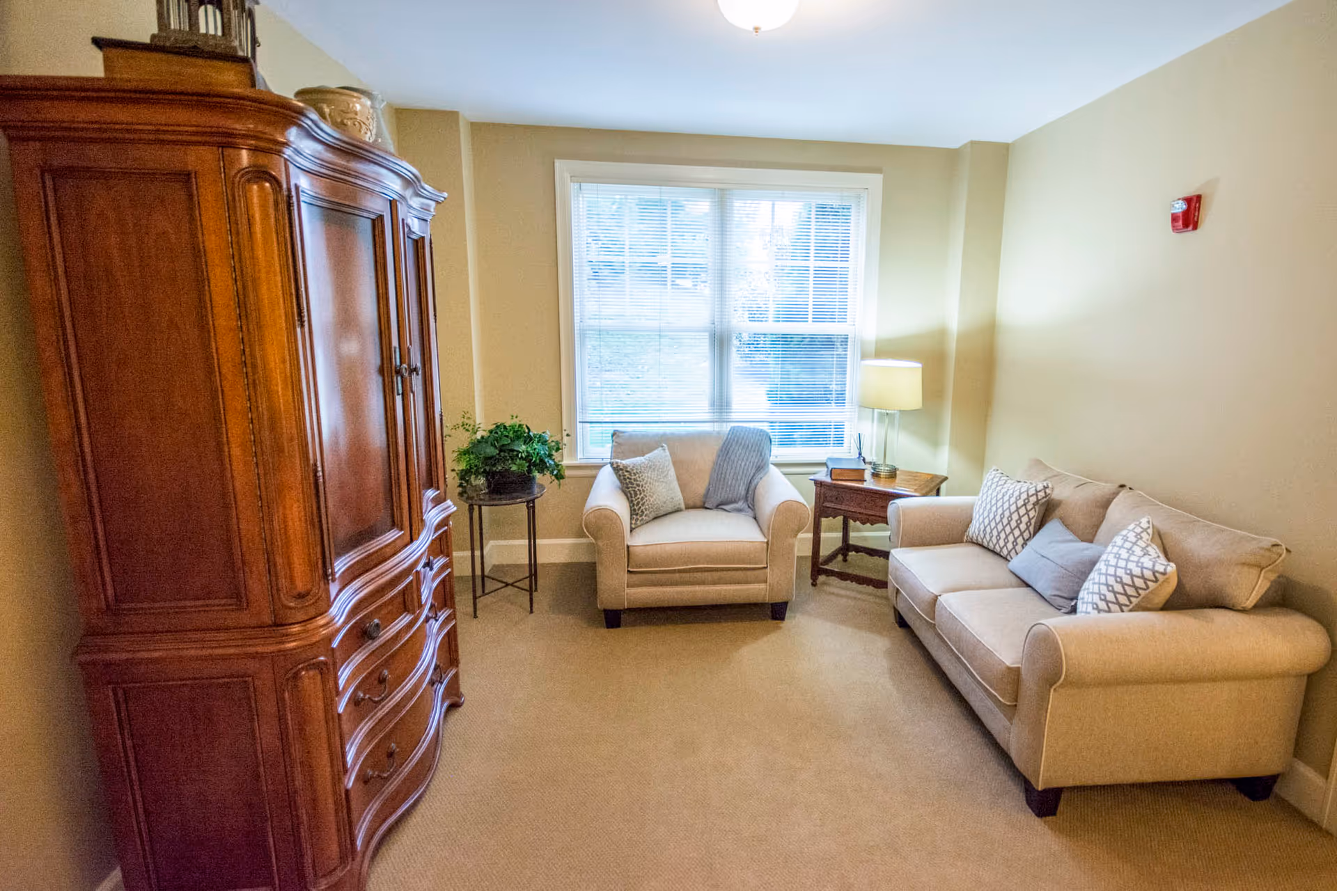 A cozy living room with beige walls and carpet, featuring a beige sofa with patterned pillows, a matching armchair with pillows and a throw, a wooden side table with a lamp, a small round table with a potted plant, and a large wooden armoire. A window with blinds lets in natural light.