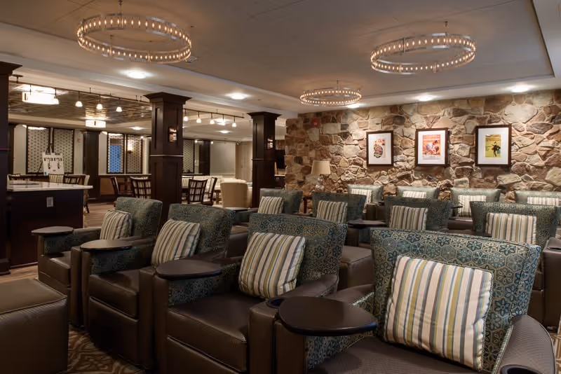 A cozy common area with multiple comfortable armchairs arranged in rows, each with patterned cushions and small attached tables. The room features a stone accent wall with three framed pictures and warm lighting from circular chandeliers on the ceiling. In the background, there are additional seating areas and wooden columns.