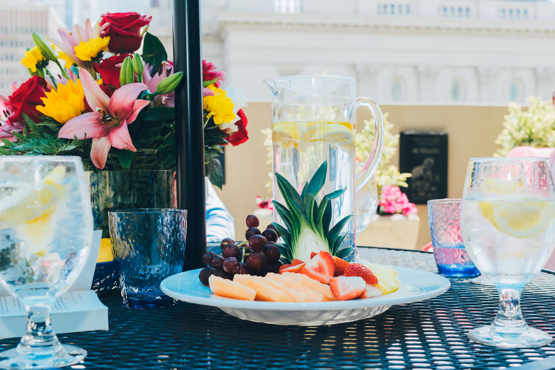 A sunlit outdoor table with a pitcher of lemon water, glasses, a plate of fresh fruit, and a vase of colorful flowers.