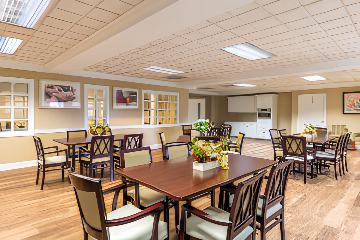 A spacious dining room with several wooden tables and chairs arranged neatly. The room has a light-colored ceiling with fluorescent lights, beige walls adorned with framed artwork, and a wooden floor. There are decorative plants placed on the tables, and a small kitchen area with white cabinets is visible in the background.