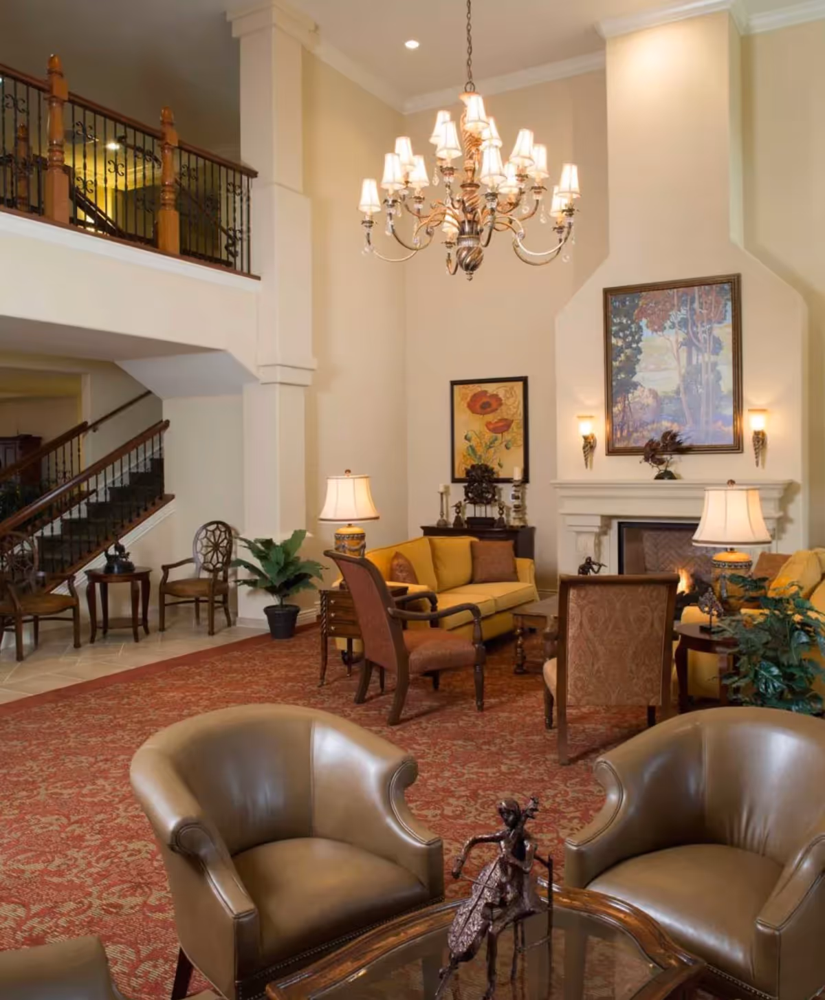 Elegant communal living room with leather armchairs, a chandelier, fireplace, and seating arranged on a red patterned carpet.
