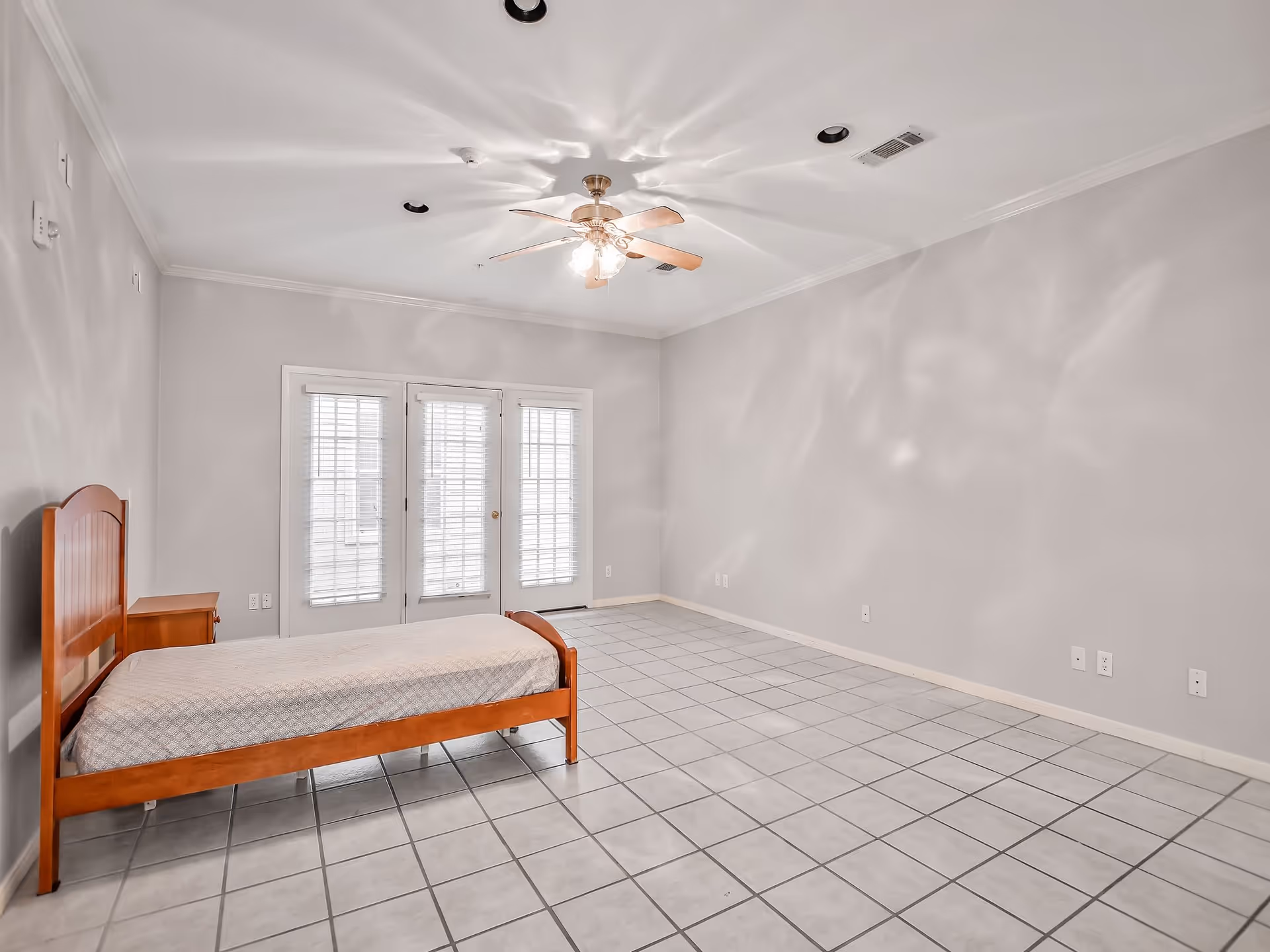 A spacious bedroom with a single wooden bed and matching nightstand on the left side. The room has light gray walls, white tiled floor, and a ceiling fan with lights. At the far end, there are three glass doors with white blinds allowing natural light to enter.