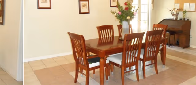 A dining area with a wooden table and six matching chairs with white cushions. There is a vase with flowers on the table. The room has tiled flooring and light-colored walls with framed pictures. In the background, there is a wooden piano with decorative items on top and a door with glass panels.