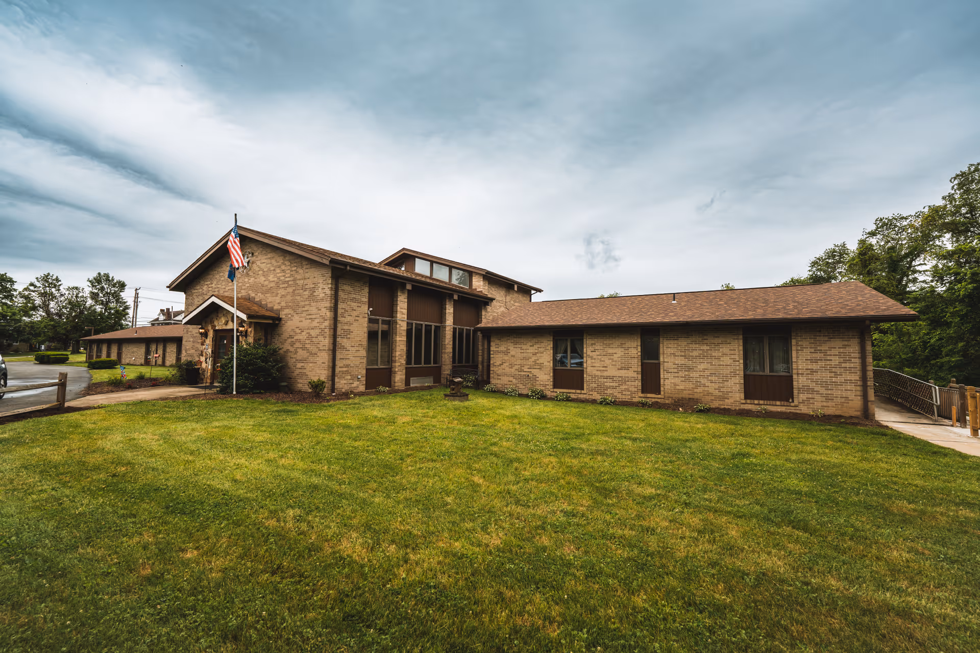 Brick senior living facility building with an American flag in front and a large grassy lawn under a cloudy sky.