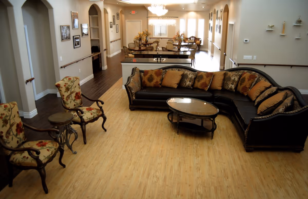 A spacious living room area in an assisted living facility featuring a large dark sectional sofa with multiple patterned cushions, a glass-top coffee table, two floral upholstered armchairs with a small round side table between them, and a dining area with tables and chairs in the background. The room has wood flooring, beige walls, framed pictures, and handrails along the walls.