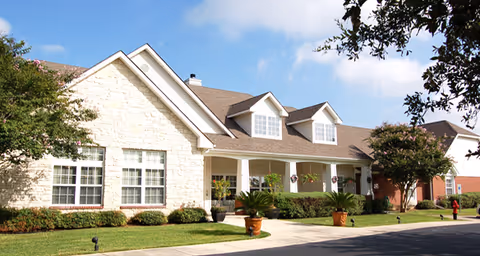 Exterior view of a single-story senior living facility building with a stone facade, multiple windows, a covered porch with columns, potted plants, and a well-maintained lawn under a partly cloudy sky.