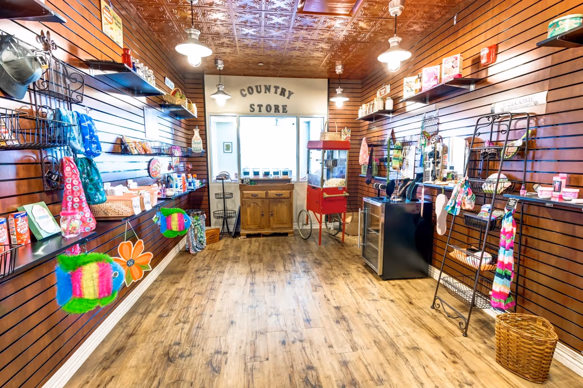 Interior view of a country store with wooden walls and floor, shelves displaying various colorful items such as bags, snacks, and decorations, a popcorn machine, and a small wooden cabinet under a window with the words 'COUNTRY STORE' above it.