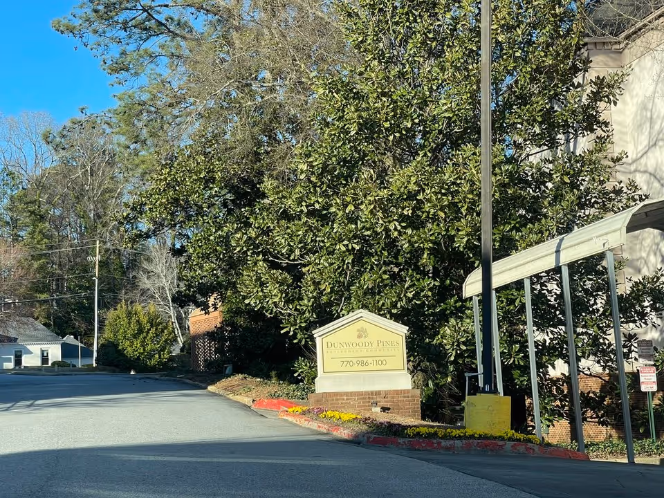 Driveway entrance with a Dunwoody Pines Retirement Community sign, landscaping, and a covered walkway beside a building.