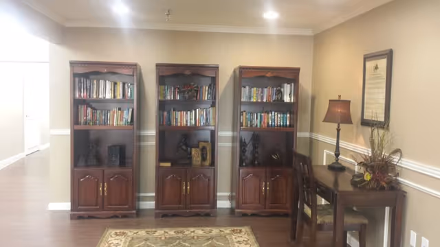 A quiet interior space featuring three wooden bookshelves filled with books and decorative items, a wooden desk with a chair, a table lamp, and a framed document on the wall. The room has beige walls with white trim and a patterned rug on the wooden floor.