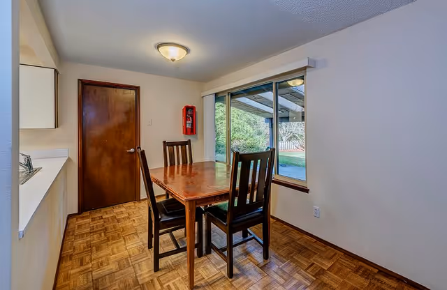 Small dining area with a wooden table and four chairs by a large window, parquet flooring, and a closed wooden door.