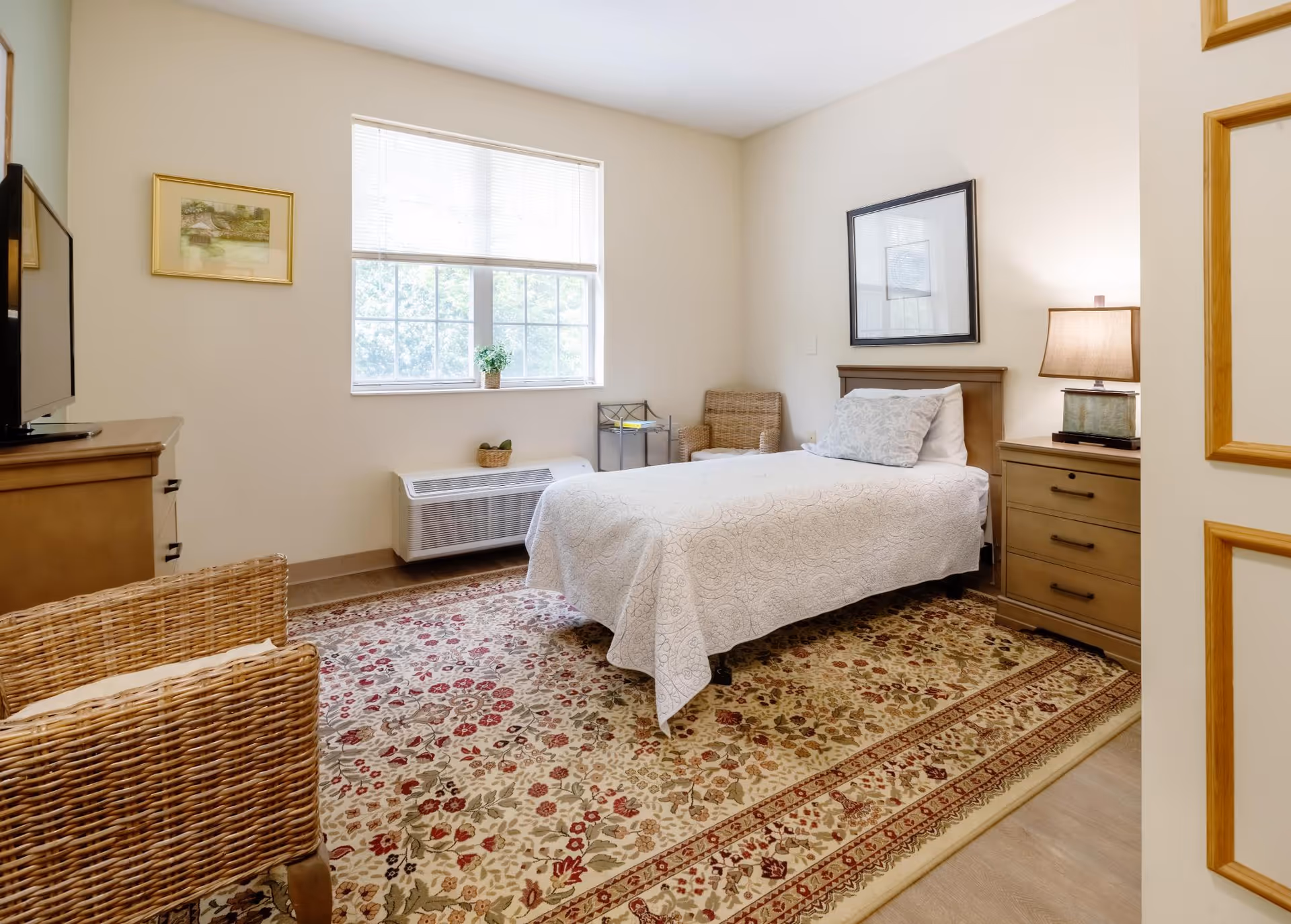 A bright and cozy bedroom with a single bed covered in a white quilt, a wooden nightstand with a lamp, a wicker chair, a dresser with a TV on top, a large window with blinds, and a decorative floral rug covering the floor.