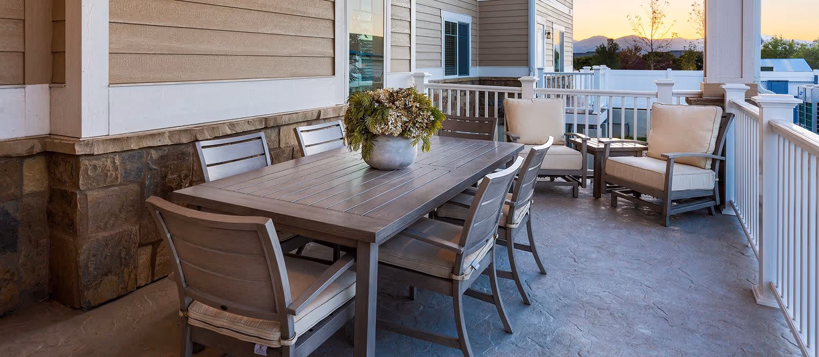 Outdoor patio area with a large rectangular wooden table surrounded by six wooden chairs with cushions. There are two cushioned armchairs and a small side table near the railing. A potted plant with flowers is placed on the table. The patio has a stone and siding exterior wall and white railing, with a view of trees and mountains in the background during sunset.