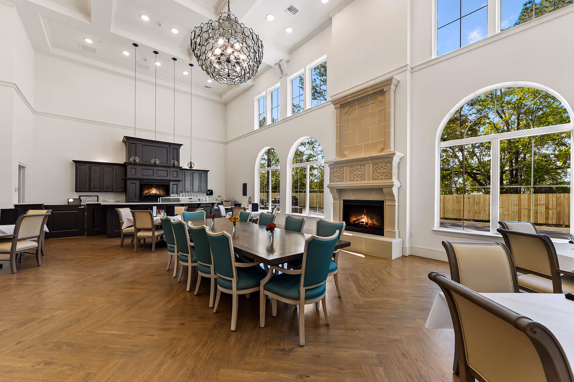 Spacious dining room with a long wooden table surrounded by teal and beige chairs, a large decorative chandelier hanging from a high ceiling, a fireplace with intricate stonework, large arched windows letting in natural light, and a dark wood cabinet area with a built-in oven and pendant lights.