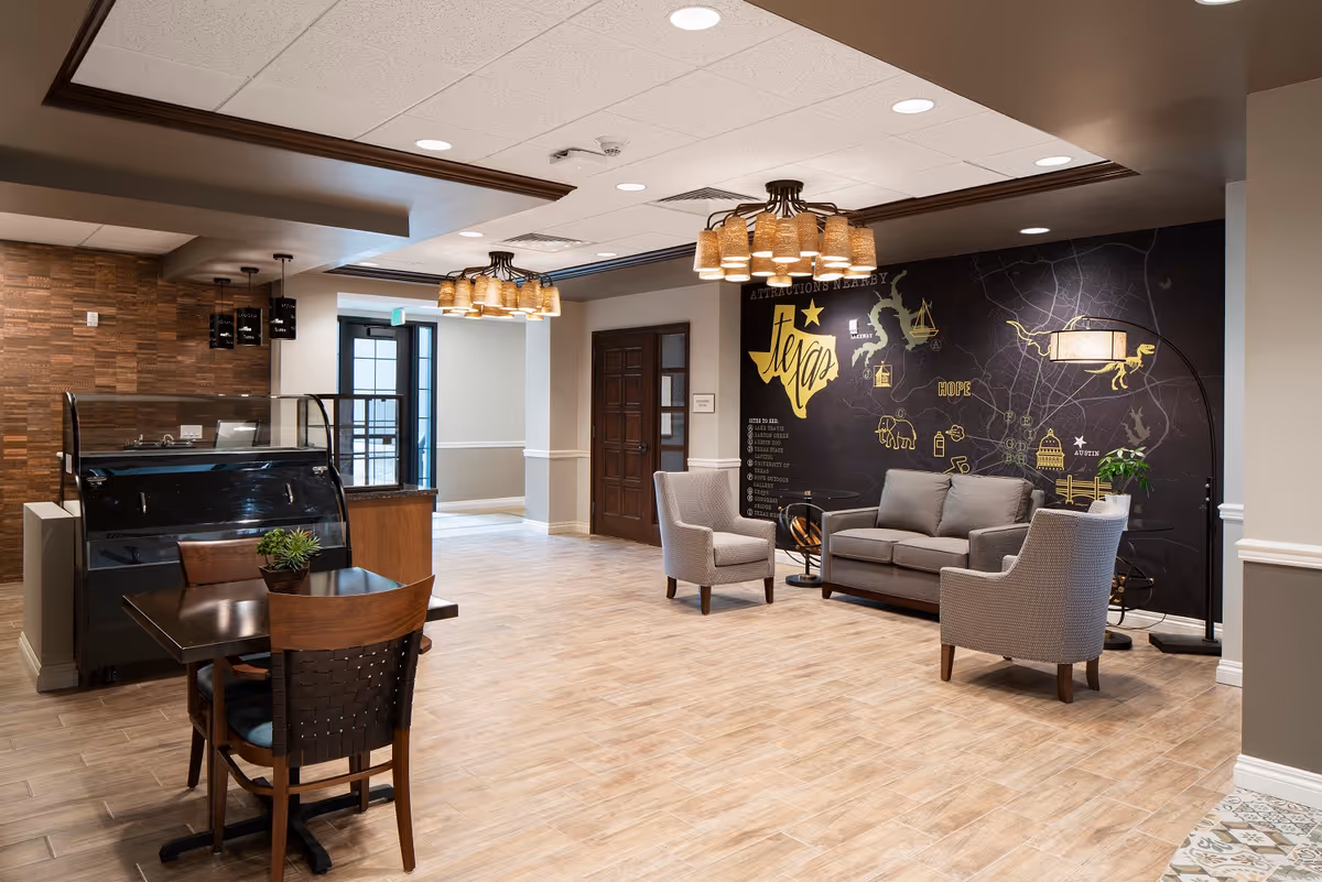 Spacious senior living lounge with armchairs, a sofa, a small dining table, and a decorative map mural on the wall.