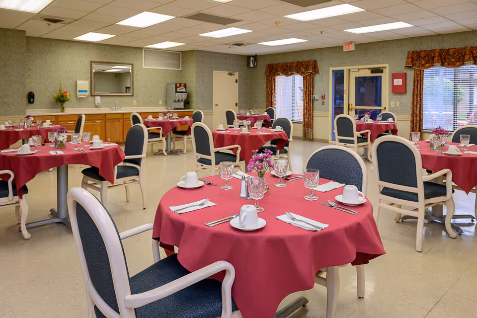 Dining room with multiple round tables covered with red tablecloths, each set with white cups, glasses, silverware, and small flower arrangements. Chairs with blue upholstery surround the tables. The room has green patterned wallpaper, a large mirror, a beverage dispenser, and windows with floral curtains.