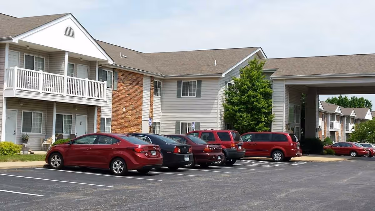 Exterior view of a senior living facility named Heritage Place showing a parking lot with several parked cars in front of a two-story building with balconies and a covered entrance.
