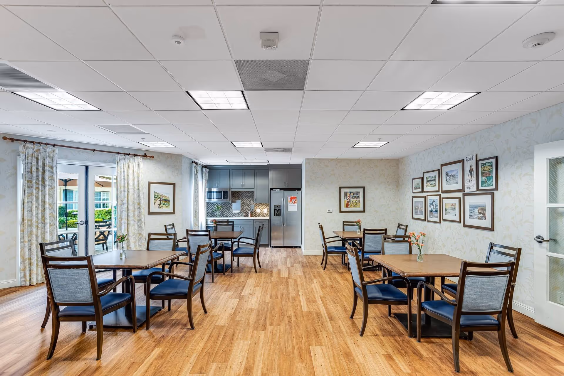 A bright dining room with wooden floors and several tables surrounded by chairs with blue cushions. The room features a small kitchen area with gray cabinets, a microwave, and a stainless steel refrigerator. There are framed pictures on the walls and large windows with patterned curtains letting in natural light. A glass door leads to an outdoor patio area.