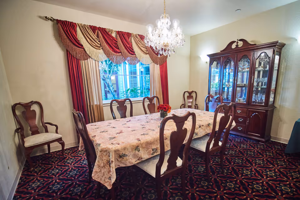 Formal dining room with a long table covered by a floral tablecloth, wooden chairs, a chandelier, draped window treatments, and a china cabinet.