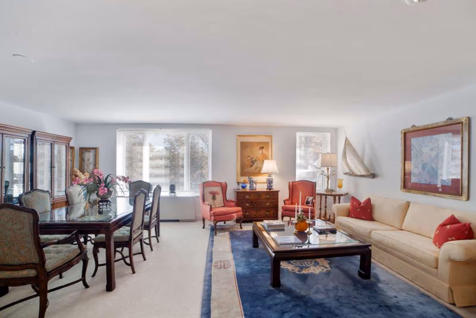 A bright and spacious living and dining area with large windows letting in natural light. The room features a beige sofa with red pillows, two red armchairs, a wooden coffee table on a blue rug, and a wooden dining table with upholstered chairs. Decorative items include framed artwork, a model sailboat on the wall, and a vase with flowers on the dining table.