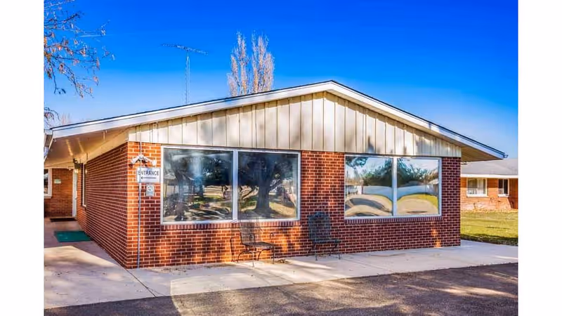 Exterior view of a single-story brick building with large windows and a sloped roof under a clear blue sky. Two chairs are placed on the sidewalk in front of the building. A sign near the entrance indicates accessibility.