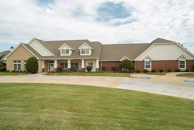 Exterior view of a single-story senior living facility building with a combination of brick and siding, a sloped roof, and a well-maintained lawn in front. There are bushes and small trees near the building, a concrete driveway, and parking spaces including handicapped spots.