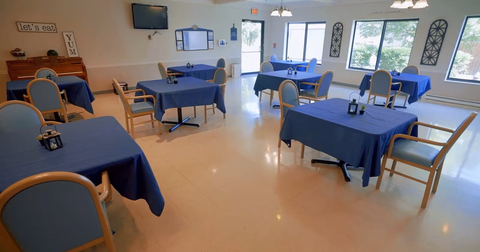 A bright dining room with several tables covered in blue tablecloths, each surrounded by light wooden chairs with blue cushions. The room has large windows letting in natural light, wall decorations including signs that say 'let's eat' and 'YUM', and a wall-mounted TV.