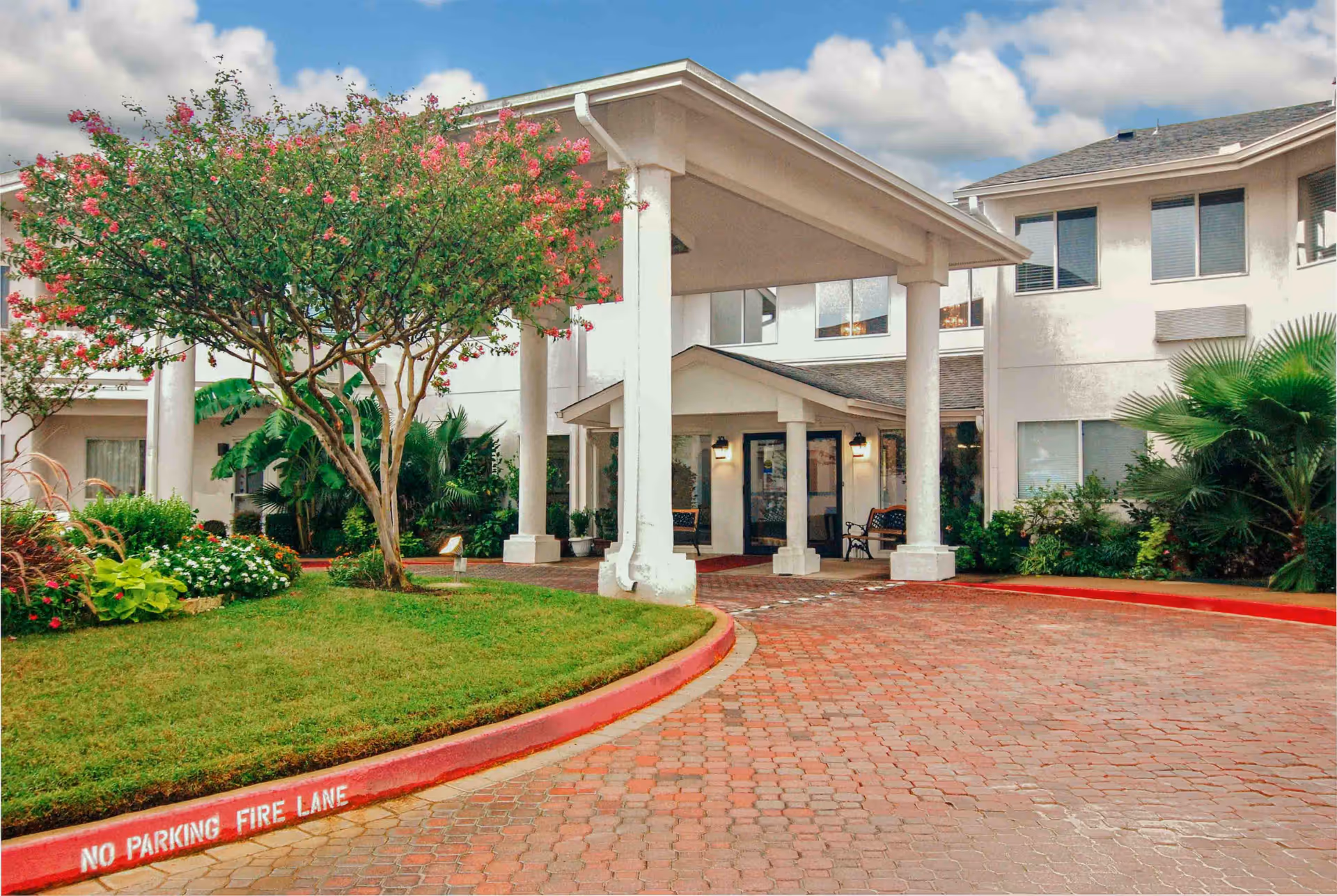 Entrance of Arlington Plaza - Sky Active Living facility showing a covered driveway with white pillars, a brick-paved circular driveway, green lawn with flowering trees and shrubs, and a two-story building with multiple windows.