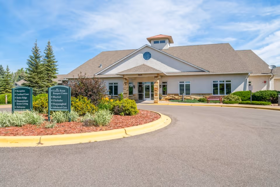 Front entrance of a single-story senior living facility with a circular driveway, landscaped beds, and directional signs.