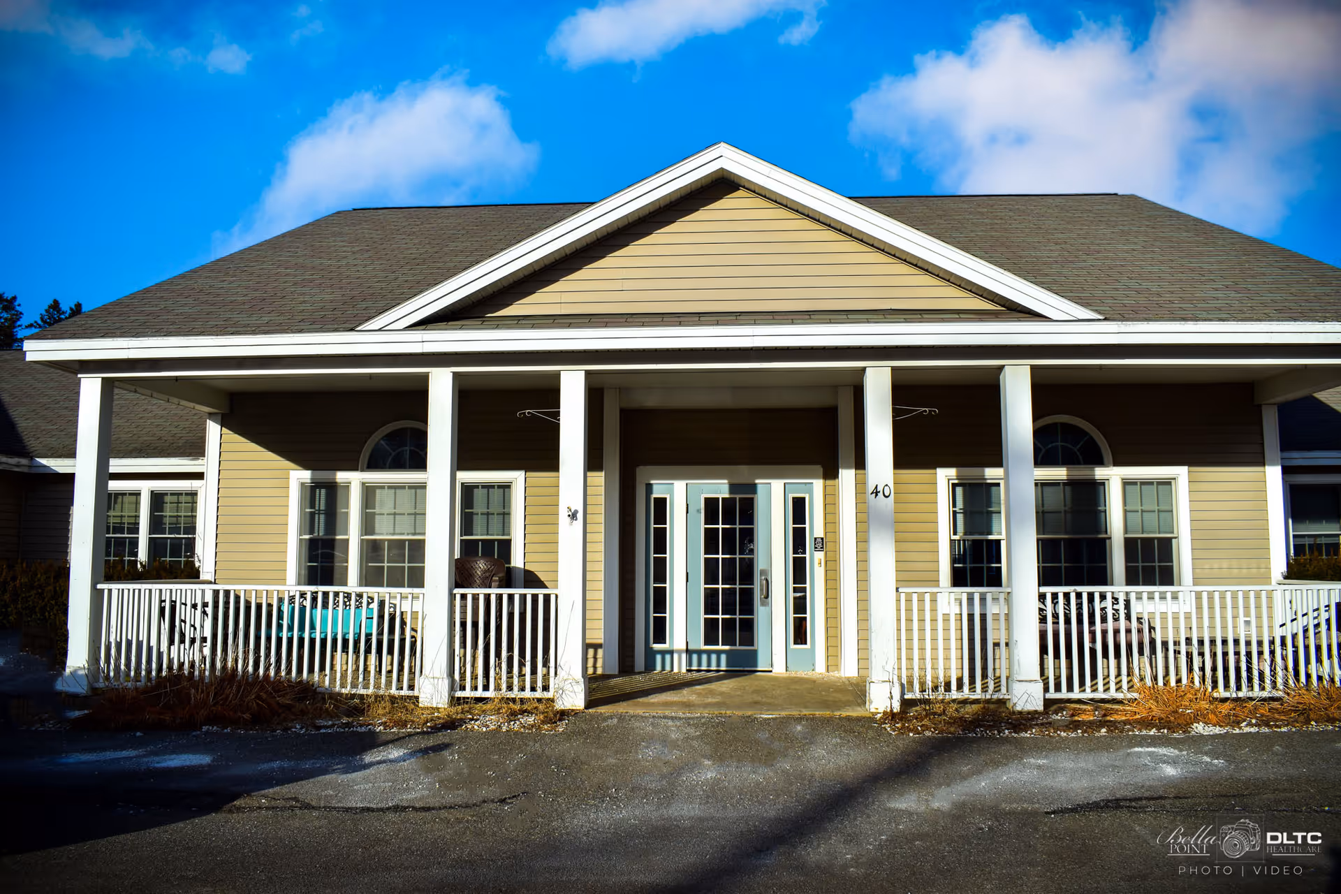 Front exterior view of a single-story building with beige siding, white trim, and a covered porch supported by white columns. The entrance features double glass doors with sidelights, and there are multiple windows on either side. The sky is blue with some clouds.