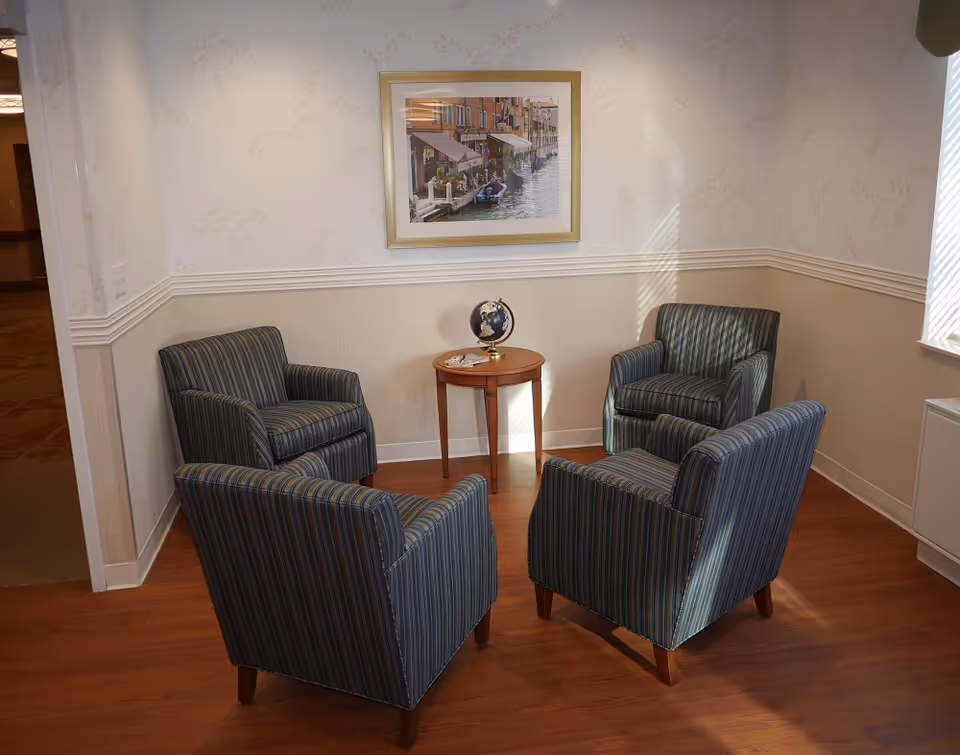 A small seating area with four striped armchairs arranged around a round wooden side table that holds a small globe and some coasters. The walls are light-colored with a chair rail and a framed picture of a canal scene hanging above the table. Sunlight streams in from a window on the right.