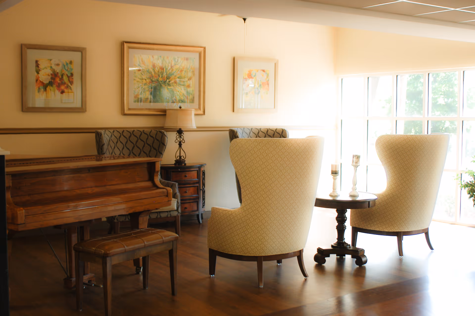 A bright sitting area with a wooden piano, upholstered armchairs around a small table, framed artwork, and large windows.