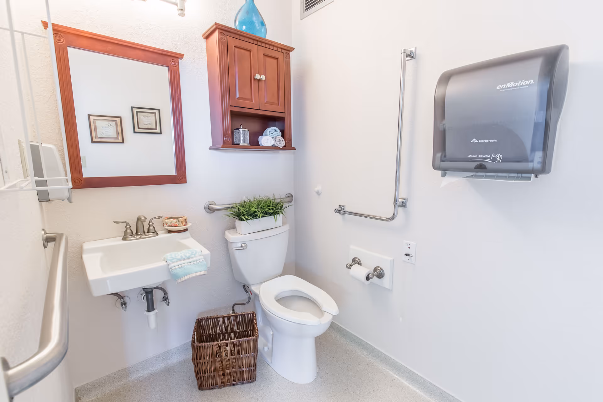 A clean and well-lit bathroom with a white toilet, a white sink with a silver faucet, a wooden framed mirror above the sink, and a wooden cabinet mounted on the wall. There is a wicker basket on the floor, a small plant on the toilet tank, and a paper towel dispenser on the wall. The bathroom has grab bars for accessibility.
