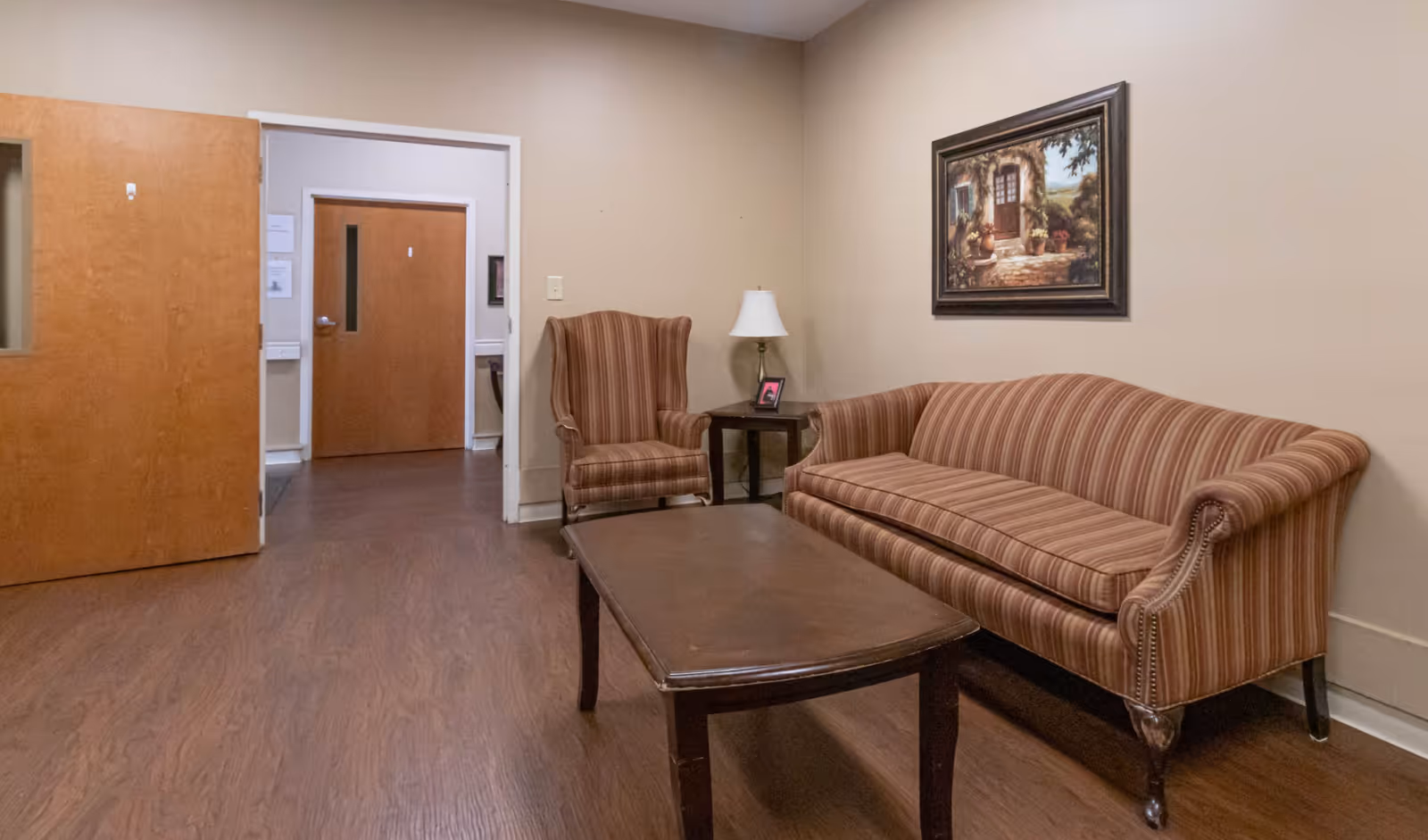Small sitting area with a striped sofa and armchair, wooden coffee table, side table with lamp, and a framed painting on the wall.