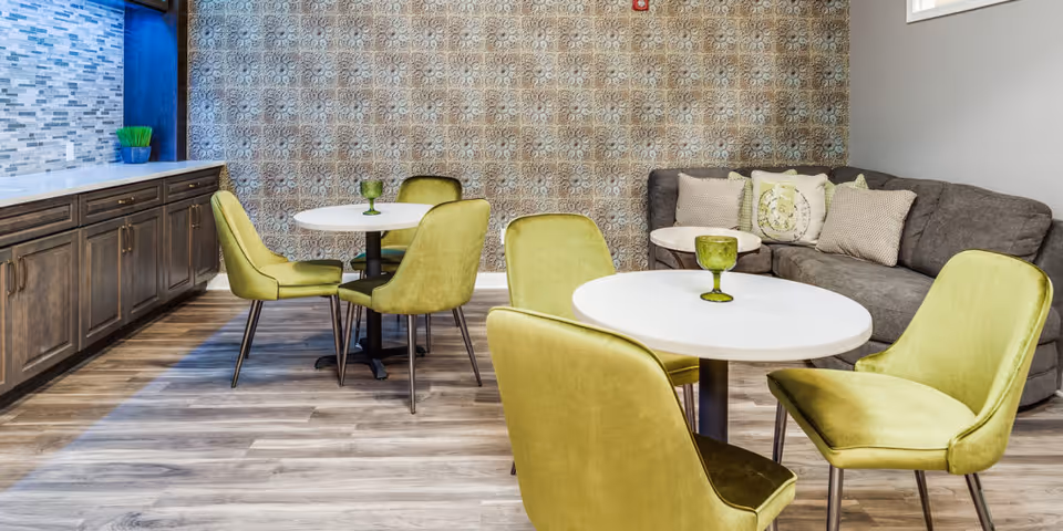 A cozy interior common area with two round white tables surrounded by green upholstered chairs. A gray couch with decorative pillows is against the wall with patterned wallpaper. There is a long wooden cabinet with a tiled backsplash and a small green plant on the left side.